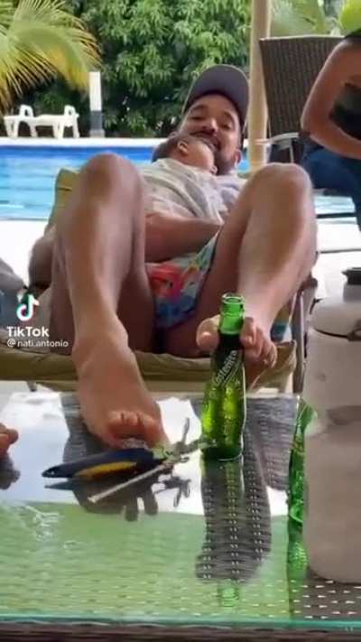 Opening a bottle with his feets while lying down holding a baby