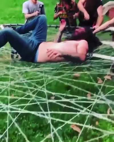 Shirtless guy jumps off step ladder onto barbed wire trampoline and gets his hair stuck