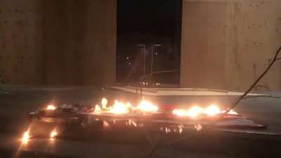 Downtown Portland at the federal courthouse, anti DHS/ICE and StopLine3 protesters gather. A few of the remaining uncovered windows have been broken. An American flag burns in front of the door. Via Garrison Davis
